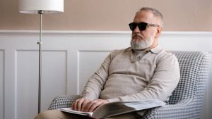 Senior man with sunglasses reading a braille book in a cozy modern interior.